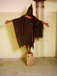 A photograph shows a person standing on a box with arms held out. The person is covered in shawl-like attire and a full hood that covers the face completely.