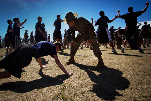 A photograph shows a person doing pushups while a military leader stands over the person; other people are doing jumping jacks in the background.